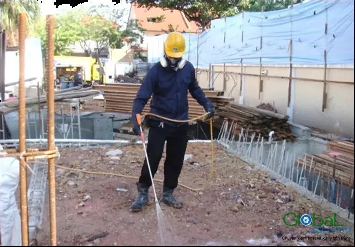 Kỹ thuật viên đội mũ bảo hộ, đeo mặt nạ phun thuốc phòng mối lên nền đất hố móng giữa các cây thép chờ