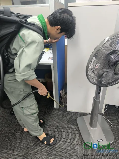 Kỹ thuật viên phun thuốc diệt gián kẽ tủ cạnh quạt đứng trong văn phòng