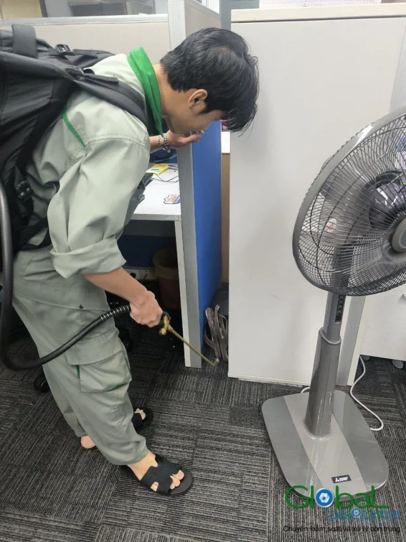 Kỹ thuật viên phun thuốc diệt gián kẽ tủ cạnh quạt đứng văn phòng