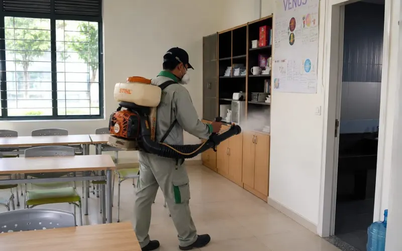 Kỹ thuật viên đeo máy phun trong phòng chức năng trường học có bàn ghế và tủ gỗ